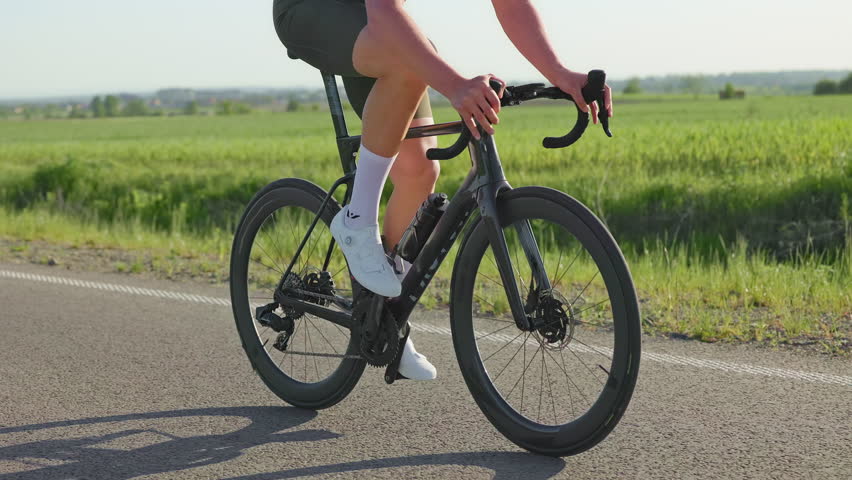Cropped view of muscular adult in sportswear using bike computer while riding racing cycle on paved road in summer. Well-built professional going up gentle slopes while exploring local rural area.