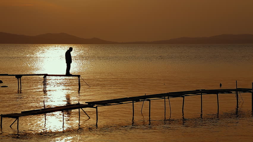 A lone fisherman stands on the pier and looks at the water at sunset.