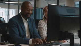 Mature man and young woman in businesswear smiling and talking by computer on desk in office