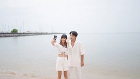 Happy Asian couple using mobile phone taking selfie together at seaside in sunny day. Man and woman enjoy and fun outdoor lifestyle travel at tropical island beach on summer holiday vacation.