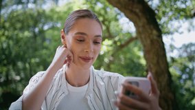 Girl talking at video chat holding smartphone under forest greenery close up. Happy attractive woman calling online using cellphone in sunny park. Cheerful lady smiling to mobile phone camera outdoors