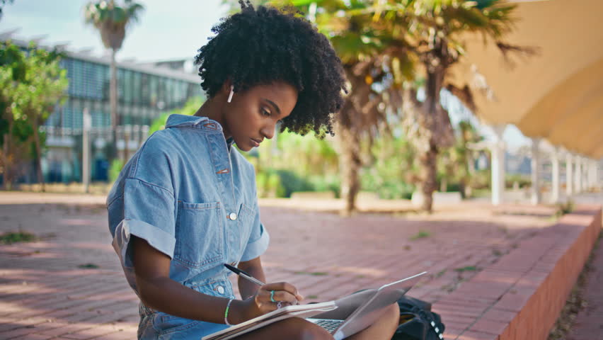 Girl learning online lesson making notes in copybook sitting sunny street close up. Smart african american university student doing homework in campus area. Curly teenager using laptop for studying. Royalty-Free Stock Footage #1107171779