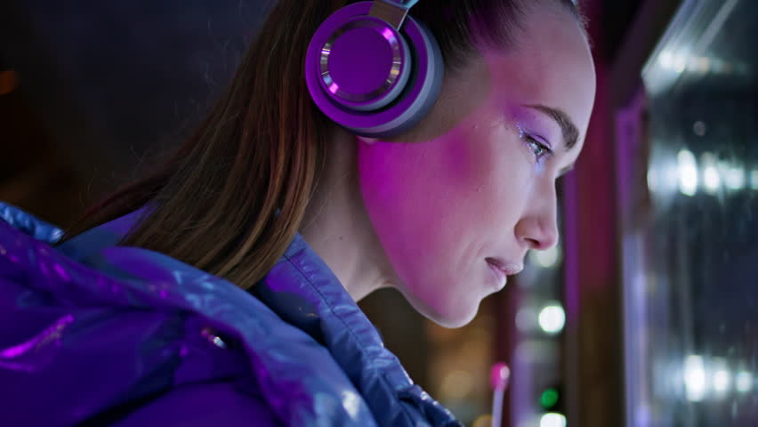 Trendy young woman looking snacks in vending machine late evening close up. Attractive girl in wireless headphones choosing food in automat standing alone at night city. Pretty brunette enjoy nightout