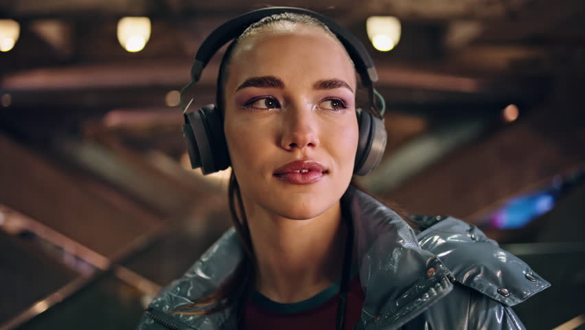 Gorgeous stylish woman wearing wireless headphones looking camera confidently close up. Portrait of beautiful teenage girl listening music in modern headset at twilight. Brunette enjoy night life. Royalty-Free Stock Footage #1107171963