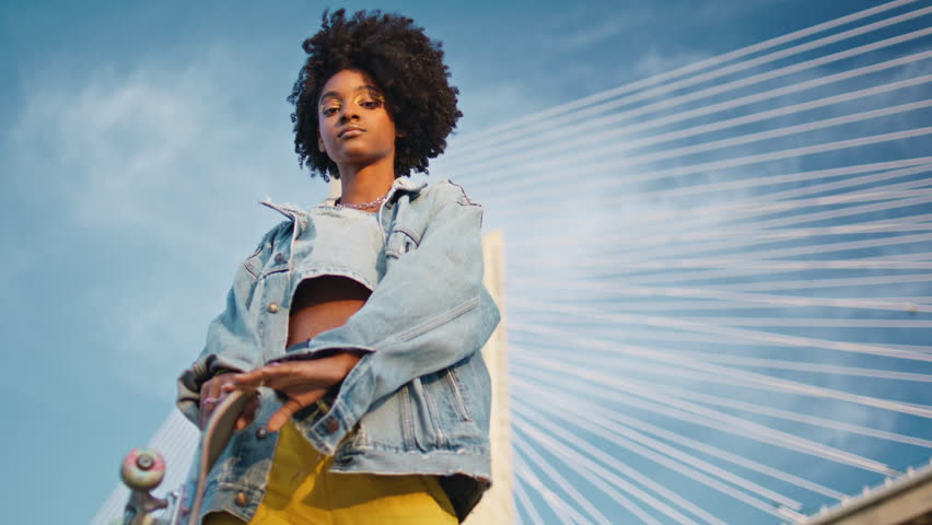 Trendy girl skateboarder looking camera in front beautiful blue sky. Attractive african american female teenager posing outdoors holding modern skate board. Curly cheeky hipster enjoy summer leisure. Royalty-Free Stock Footage #1107171995