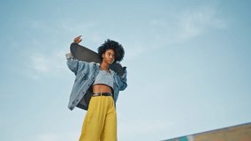 African american skater walking street with modern longboard on shoulders. Cool beautiful girl posing in front blue sky holding skateboard. Trendy curly female skateboarder looking camera confidently.