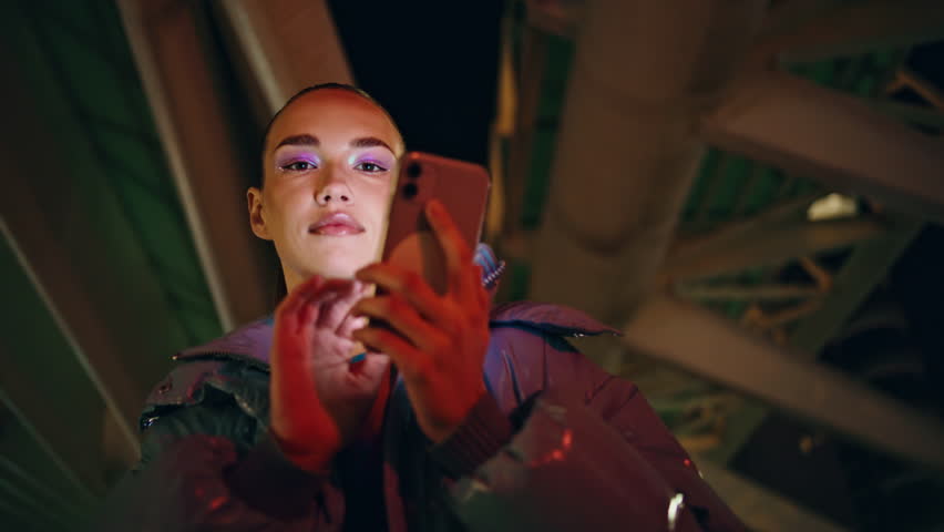 Trendy young girl typing phone at night standing in front modern urban construction closeup. Serious attractive woman using smartphone to search information Internet surfing. Technology in life