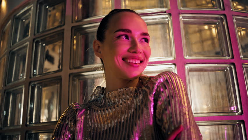 Luxury young woman smiling to camera standing in front glass wall night club close up. Portrait of trendy cheerful party girl in sparkling clothes posing at evening. Attractive model with happy smile