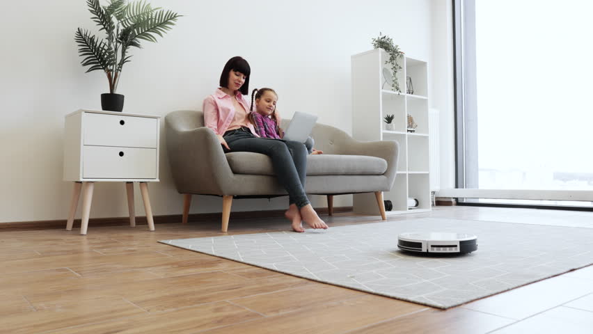 Pretty mother and daughter resting on sofa with computer while robot cleaner handling debris on floor. Happy caucasian ladies scrolling digital photo album on portable gadget with smart home device.