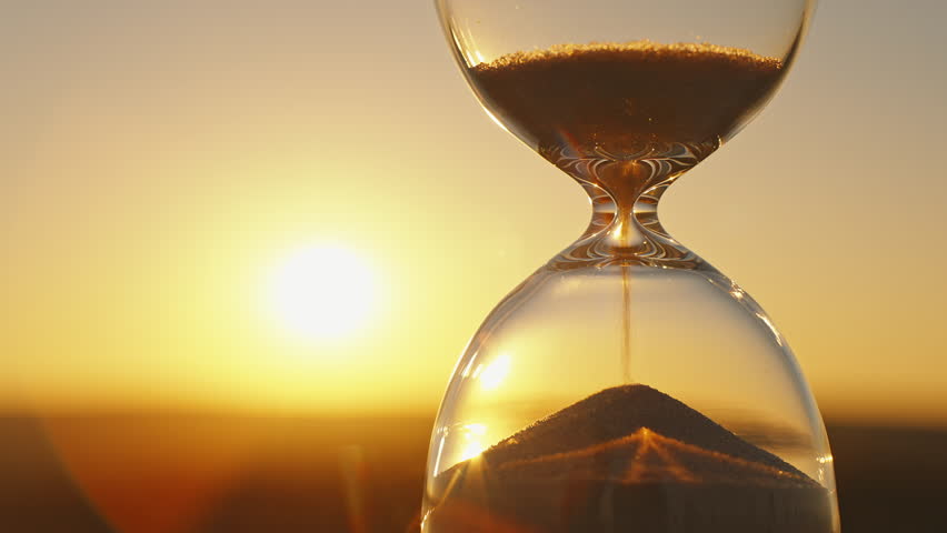Hourglass concept time on blurred background of golden sun disk sky in sunset rays in summer close-up. Golden sand pours quickly. Time concept sand. Relax Royalty-Free Stock Footage #1107175323