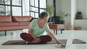 Old woman make yoga exercise at home. Mature woman have meditation. She look at video on laptop online courses