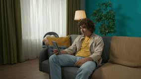 Young handsome man in casual clothing, sitting on the sofa in the living room with tablet and having a conversation by a video call
