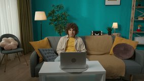 Young man in casual clothing, sitting on the sofa in the living room talking by video call on laptop