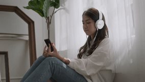 Happy young asian woman relaxing at home.Female smile sitting on floor and holding mobile smartphone.Listening to music on lazy and relax day at summer or rainy season