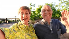 Senior couple looking to camera smiling, having video call, talking to friends outdoors. Portrait of cheerful smiling retired man and woman have fun together at summer sunset. Enjoy romantic holiday