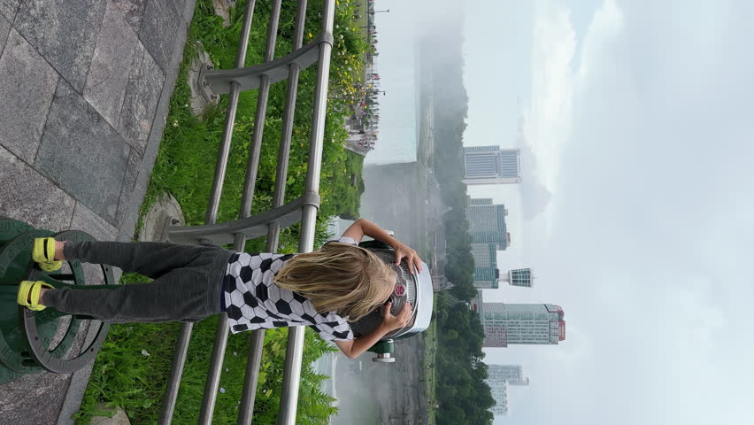 6 years old caucasian boy looks into touristic binoculars to see the Niagara river and the Niagara falls better. Canadian Ontario is on the other bank of the river, view of buildings and green trees