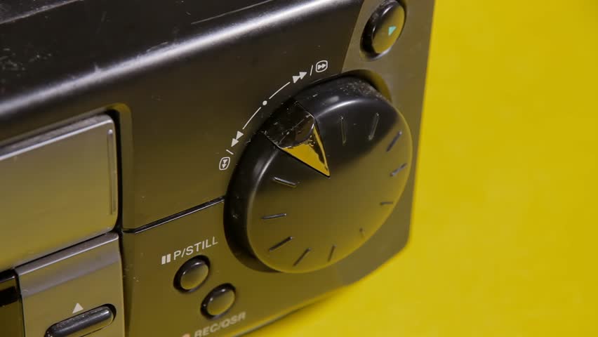 Angled close-up shot: a hand manipulates the knob of a vintage VHS VCR, commanding the videotape motor to perform various actions such as fast forward and rewind. Royalty-Free Stock Footage #1107189477