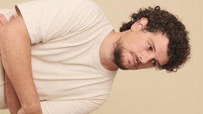 Vertical video, Thoughtful guy with curly hair dressed in beige t-shirt, with his hands folded unable to make up his mind in studio on beige background in the studio