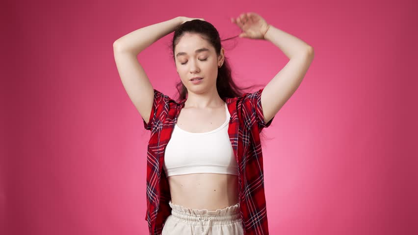 Casually dressed fit young lady adjusting long dark hair into a ponytail with hairpiece while looking relaxed and self confident against pink background | Shutterstock HD Video #1107209879
