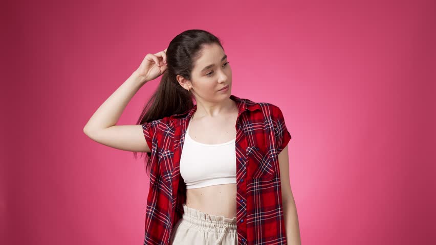 Young woman wearing casual dress letting loose her long dark hair by taking out black hairpiece while looking coquettish and confident against pink background | Shutterstock HD Video #1107209883