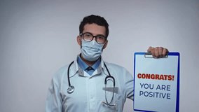 Young doctor in white coat and protective mask with stethoscope shows paper holder with text CONGRATS. YOU ARE POSITIVE and points on it.