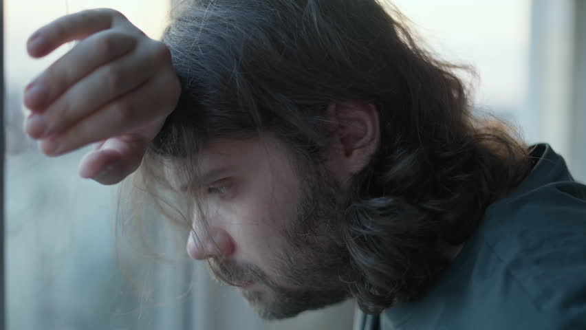 Depressed man staying alone near window with stressed problem face, closed eyes. Pensive, thoughtful. Denial, Bargaining emotions. Mental health, man depression, despair concept. Long hair, beard | Shutterstock HD Video #1107215957