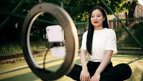 A beautiful young asian woman blogger sits on a sports field and broadcasts live on a social network. The girl is blogging, reviewing products and leaving honest feedback