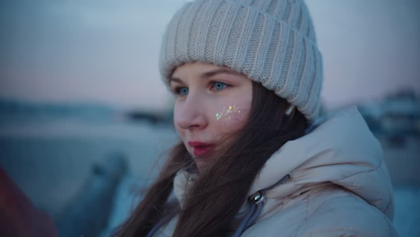 A young caucasian girl with red lipstick, blue eyes and glitter on her face in a winter jacket, long hair and a white hat takes pictures of winter sea and hills on smartphone with multi-colored case