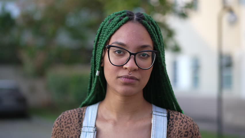 One young black woman standing outside looking at camera with serious expression. A Brazilian female person with box braids hairstyle