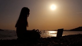 Teen look meditation coaching videos. A pretty child silhouette in lotus pose watch yoga coaching video on her notebook on seashore during nightfall time.