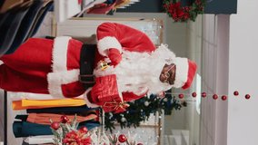Vertical video African american man dressed as Santa Claus in Christmas themed shopping mall clothing store. Employee jingling xmas bells and holding red package present in festive decorated shop