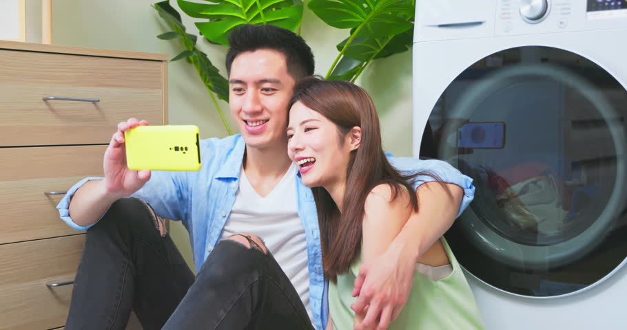 Cuddling asian couple is looking at phone together while the washing machine is cleaning clothes | Shutterstock HD Video #1107234275