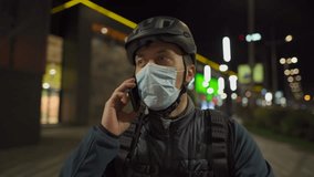 Cyclist in helmet and protective mask commuter with bicycle on way home from work talking on phone at night city. Deliveryman on bike, delivery parcels, make phone call during coronavirus quarantine. - Powered by Shutterstock - Get 15% off with code: PIKWIZARD15