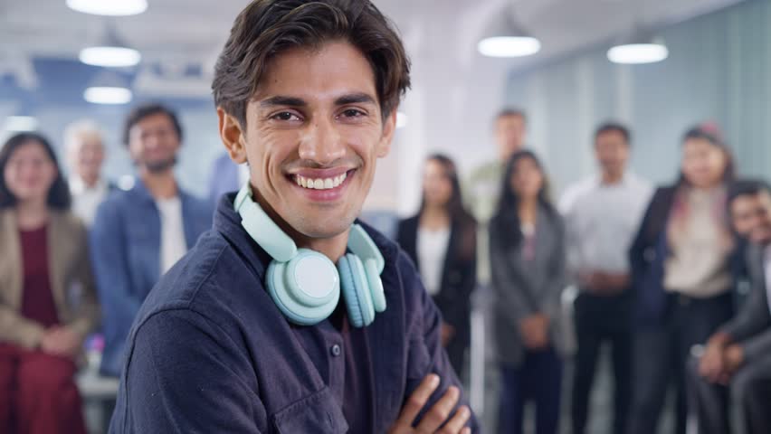 An Indian Asian happy modern male software engineer or young corporate man wearing headphones around his neck looking at the camera and smiling with a team of office employees standing in the back | Shutterstock HD Video #1107238591