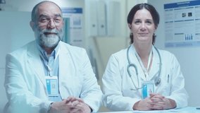 Senior male doctor and his female colleague sitting together at desk and smiling on camera during workday in clinic, both wearing white coats and stethoscopes. Medium shot, video portrait
