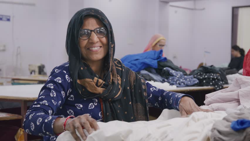 A happy middle aged Indian Asian female or woman worker in traditional or ethnic wear is interacting and working with the clothes smiling in a textile workshop or industrial firm or factory  Royalty-Free Stock Footage #1107239625
