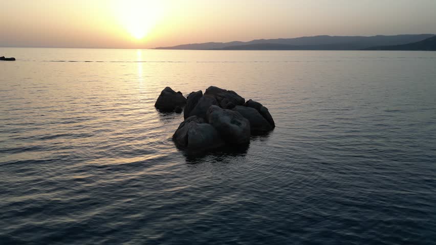Aerial drone footage circling around a rock located in the middle of transparent water during a sunset near Portogliolo Beach in Corsica