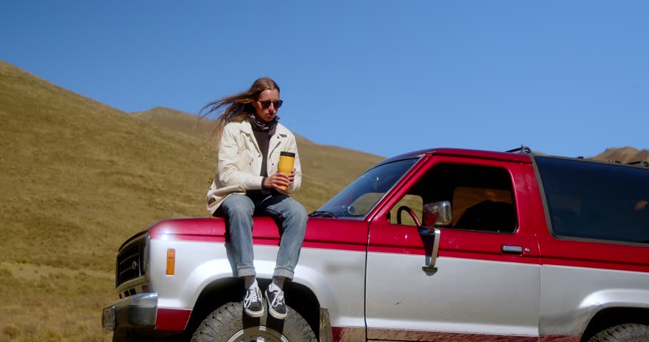 Attractive long-haired girl drinking a drink from a thermos of thermo mugs while standing near a vintage car against the backdrop of a mountain landscape. High quality 4k footage