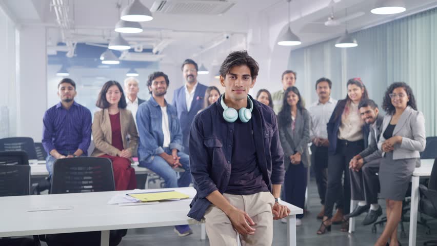 A young Indian Asian happy modern male entrepreneur or IT Professional man wearing headphones around his neck looking at the camera and smiling with a group of office colleagues standing in the back | Shutterstock HD Video #1107250331