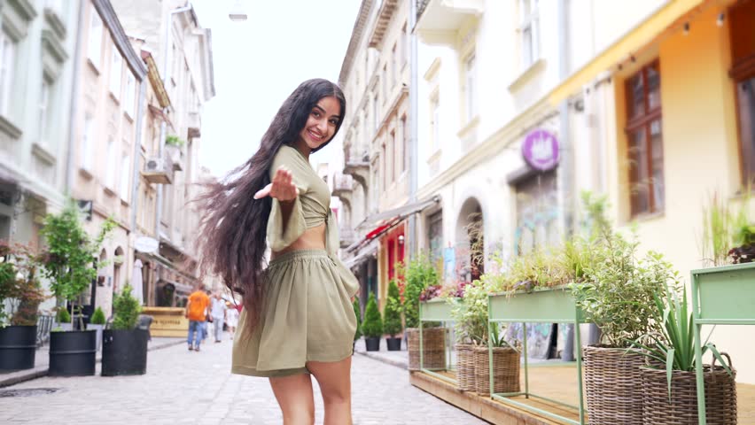 Beautiful young indian woman makes a beckoning gesture with one finger, inviting to approach, looks playfully flirting. Gorgeous girl waving hand to come with me on city street. Come here, follow me