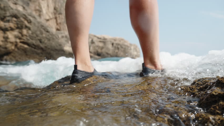 Low-angle shot capturing the view of female feet being covered by the waves of the sea as she stands on a rock at the shore, in slow motion.