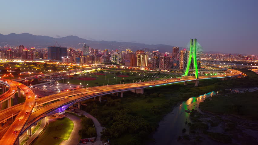 Aerial hyperlapse of cable-stayed New Taipei Bridge across Tamsui River, with busy traffic under the pylon, which is colorfully illuminated at night, and 101 Tower in background in Taipei City, Taiwan