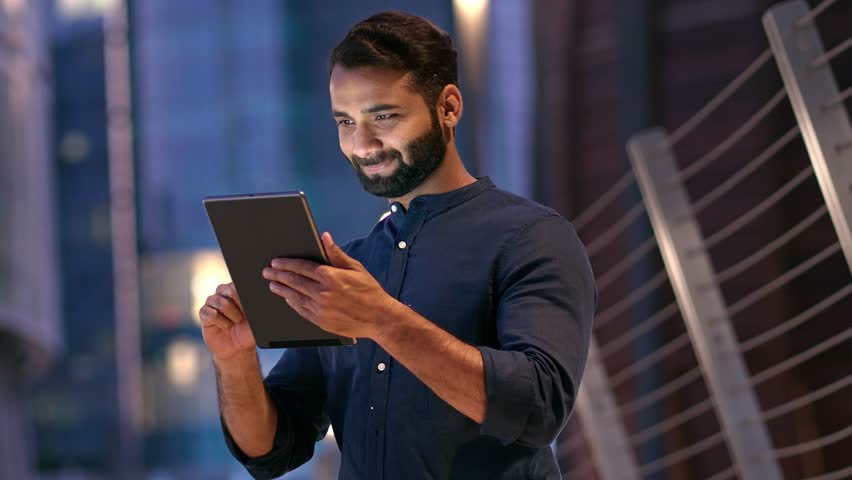Smiling young indian business man professional, eastern businessman executive standing outdoors on street reading ebook holding using digital tablet online technology in night city with urbans lights. Royalty-Free Stock Footage #1107255867
