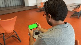 Adult man in his 30s with glasses looking at a green screen on his cell phone in a classroom. Watching on mobile phone. Ready chroma key for mock up. Horizontal screen. 