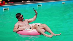 Relaxed Caucasian sportive man swimming in the pool. Man sits in the inflatable donut having a video chat on his phone.