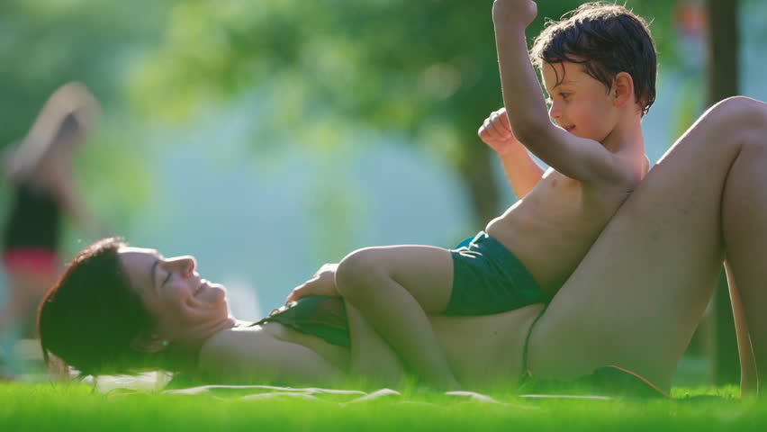 Mother and child laid on grass by poolside during summer day, affectionate candid loving family moment, motherhood lifestyle moment bonding her son