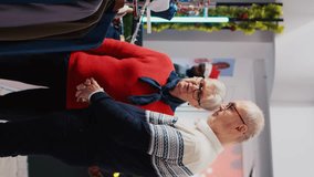 Vertical video Senior couple browsing through clothes racks in festive adorn clothing store during Christmas shopping spree. Old clients planning to purchase formal attire, enjoying holidays