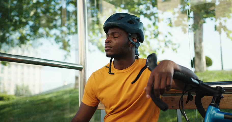 Positive African American man sits at bus stop with hand resting on bicycle handlebar. Smiling man in helmet relaxes from active pastime slow motion