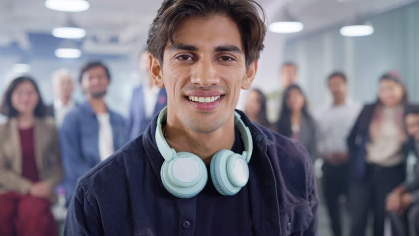 An Indian Asian happy modern tech savvy male or man wearing headphones around his neck looking at the camera and smiling with office employees standing in the back in a corporate start-up business | Shutterstock HD Video #1107279557