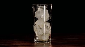 pouring orange juice into a clear glass full of ice qubes and a black background behind it with a wooden table